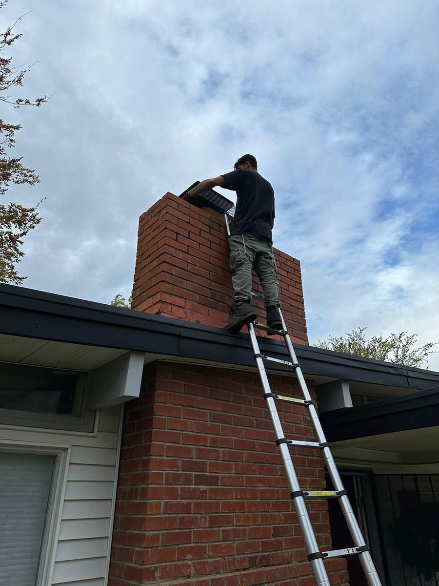 Best Chimney LLC technician on a telescoping ladder installing a mesh chimney cap on a red brick chimney