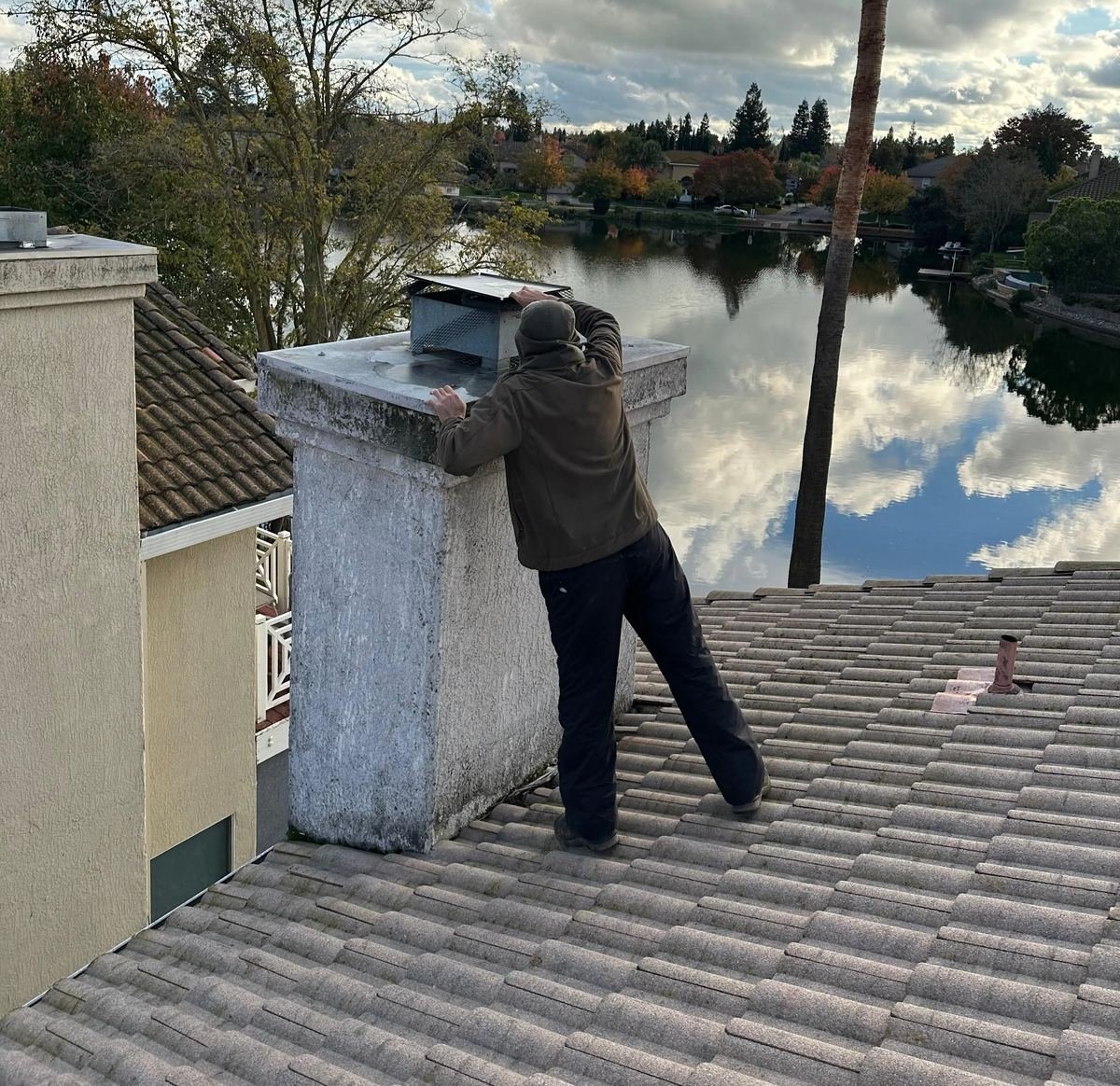 Best Chimney LLC technician fitting a metal chimney cap and chase top to a stucco chimney on a tile roof overlooking a Sacramento-area lake